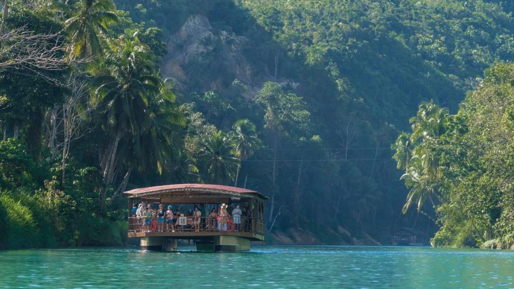 Loboc River cruise boat carrying tourists through turquoise water with lush palm‑covered riverbanks and forested hills in the background