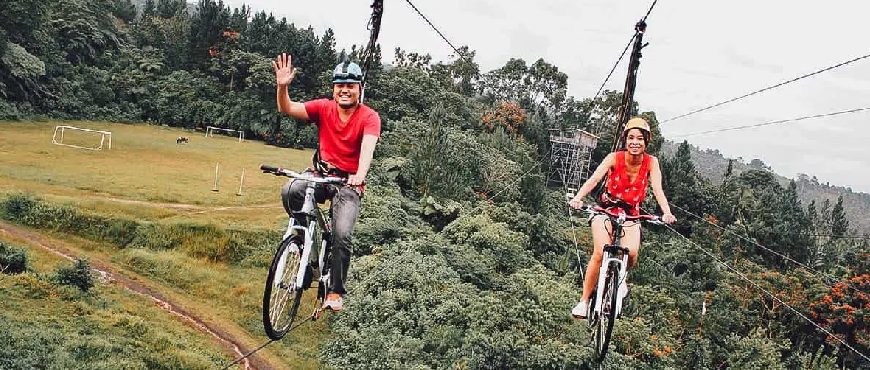 Two riders on a bicycle zipline in Bohol, Philippines, crossing above green fields and forest scenery